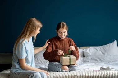 Mutlu genç kız kız yatakta oturuyor paketlenmiş hediye kutusunu açıyor, annesinden hediye alıyor. Sevgi dolu bir anne çocuğunu evde doğum günüyle kutluyor, aile özel bir olayı kutluyor.