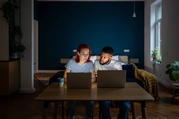 自宅で小さなオンラインビジネスを実行している若い多様な起業家のカップル 夕方に一緒にノートパソコンで作業テーブルに座って多人種の家族の男性と女性 起業家精神 — ストック写真