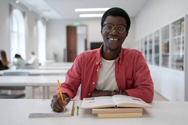 Позитивный Афроамериканец Учится Библиотеке Делает Заметки Блокноте Улыбается Глядя Камеру — стоковое фото
