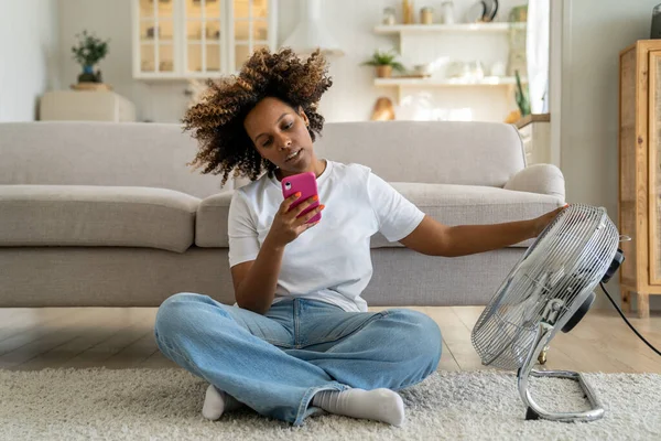 African American girl sitting on floor in living room near electric fan and using smartphone, unhappy black woman cooling down, trying to stay cool without air conditioning, suffering from heat
