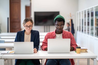 Uluslararası üniversitedeki ciddi çok ırklı öğrenciler kampüs kütüphanesindeki dizüstü bilgisayarlarda çalışıyor. Afrika kökenli Amerikalı erkek ve beyaz kadın araştırma projesinde birlikte çalışıyorlar..