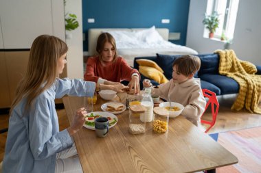 Sabahları çocukları olan bekar bir anne yemek yiyor. Annesi ve genç kız kardeşi kahve isterken elinde kaşık kakao tutan mutlu çocuk. Aile, kahvaltıda sağlıklı mısır gevreği, okuldan önce sandviç, iş.