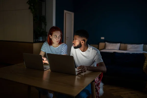 Serious multiethnic freelancers guy girl working on laptops in dark sitting at desk at home in small apartment studio. Man and woman talking, discuss job looking face to face. Work together concept.
