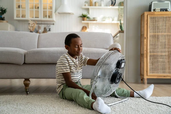 リビングルームに一人で座っている小さなアフリカ系アメリカ人の子供は 電気ファンから吹く新鮮な冷たい空気をお楽しみください 家で暑さに苦しんでいる小さな子供は 暑い中で冷えています 子供と熱波 — ストック写真