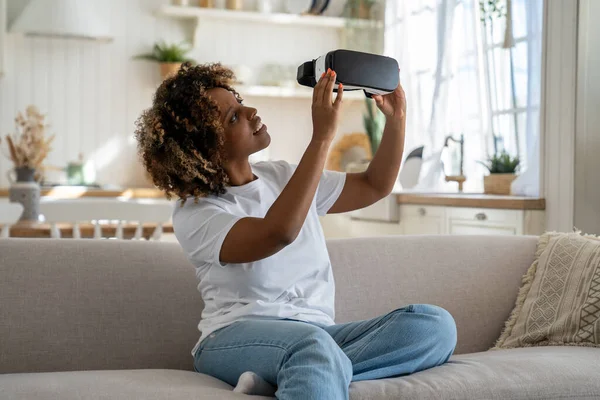 兴奋的非洲裔美国女人坐在客厅的沙发上 看着她的第一个Vr耳机 兴奋地沉浸在其中 好奇的黑人女性游戏玩家手持未来主义虚拟现实眼镜 — 图库照片