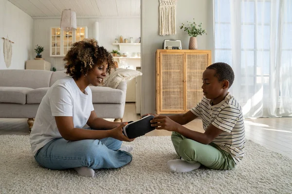 Modern African American family mother and little boy son sitting on floor with VR glasses. Mom showing gaming device 3d glasses to child at home. Immersive virtual reality and child development