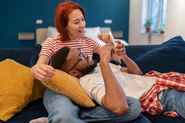 多様な幸せなカップルの男性と女性の電話のリビングルームでソファの上に横たわって見てください レジャーオンラインスマートフォンでサーフィン 携帯電話で一緒に面白いコンテンツを見てソーシャルメディアで閲覧 — ストック写真