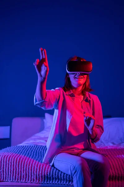 Futuristic image of young amazed millennial girl wearing metaverse VR headset touching objects in virtual world, sitting on bed under red neon light, excited woman experiencing virtual reality at home