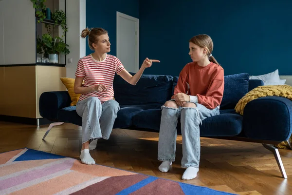 Parent-teen conflict. Strict mother scolding lecturing upset teenage girl daughter at home, pointing finger. Mom disciplining teenager while sitting together on sofa. Parenting adolescent