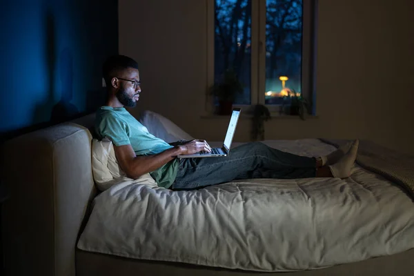 Black student guy lying in bed with laptop in evening, studying until late hours. African American remote employee working overtime from home, doing freelance work at night