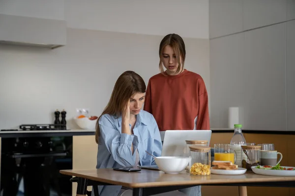 Upset depressed young woman single mother living with teen daughter sitting at kitchen table looking at laptop cant find job, mom parent struggling to pay bills, family having financial problems