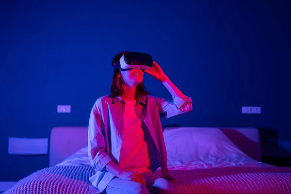 Relaxed smiling young woman in vr glasses is playing video games in dark room with purple light. Happy girl in VR goggles having fun sitting in bed. Leisure, recreation, digital entertainment concept.