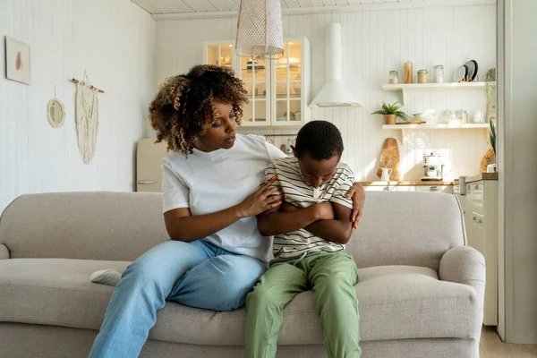 stock image Loving african american mother embracing hugging sad little son supporting child, mom parent trying to make peace with offended upset kid, sitting together on sofa at home. Parent-child conflict