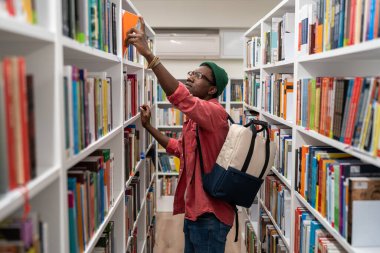 İnek öğrenci Afrikalı Amerikalı bir adam kütüphanede kitap seçiyor ve kitaplıktan alıyor. Sırt çantalı, üniversitede okuyan, hasretli bir hippi. Eğitim, yeni meslek vermek, öğrenim kavramı.