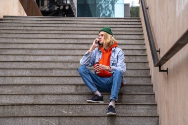 Elinde kahve fincanı olan şık bir adam merdivenlerde oturup şehir caddesinde akıllı telefondan konuşur. Şehir merkezinde uzun bir yürüyüşten sonra dinlenen adam. Modern insan dostu şehir ortamı, temiz kamusal alanlar konsepti. 