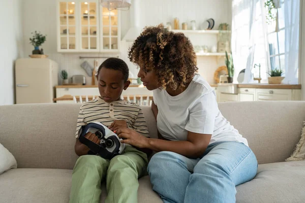 stock image Modern American family mother and little boy son sitting on sofa with new device VR headset, mom exploring virtual reality technology with child. Young woman stepping into virtual world as parent