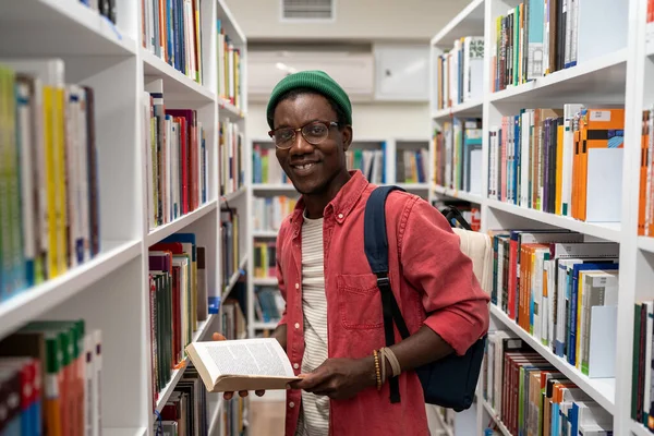 本棚の間に立っている大学図書館で本を持っているアフリカ系アメリカ人の学生を笑顔 キャンパス生活 自己啓発読書学習の概念研究を学ぶ本 — ストック写真