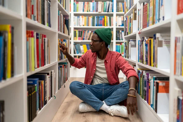 本の棚からそれを取って大学図書館で研究教科書を選択し アフリカ系アメリカ人の学生 公共の場所で勉強床に座って考えて男ヒップスター 教育学 — ストック写真