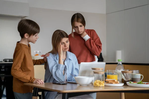 Mom with headache works laptop on kitchen with children standing near looking at screen. Tired, exhausted working woman doing distant job at home raising son daughter. Motherhood and career concept.