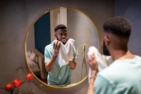 満足のいくアフリカ系アメリカ人男性の笑顔鏡の中に見える白いバスタオルは仕事の前に朝をお楽しみ保持します 自宅で気分の良いバスルームに立って幸せな黒人男性 男性の顔のスキンケアルーチン — ストック写真