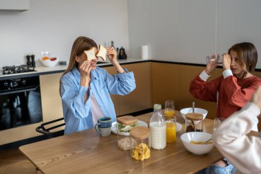 Mutlu gülümseyen kadın anne tost ekmeğiyle göz göze gelir sabah çocuklarla mutfak masasında eğlenir. Mutlu genç kız kahvaltı saatinde neşeli anne ve küçük kardeşle oynaşıyor.