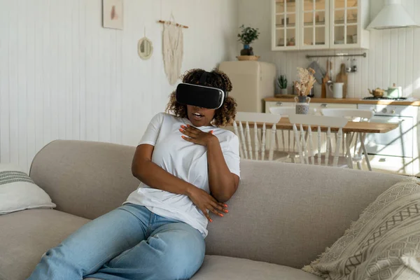 Excited wondered african american woman virtual glasses watching movie playing video games. Smiling female looks at virtual world in VR helmet sits on couch at home. Cyberspace, entertainment concept.