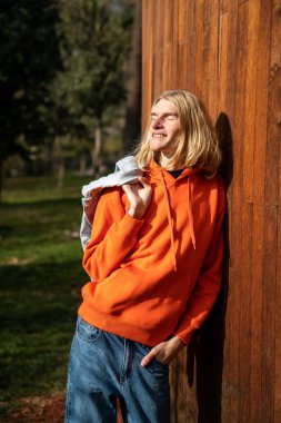 Relaxed blonde guy hipster enjoying basking sun standing near wooden fence in park with closed eyes. Slim happy man walking outdoors on nature. Rest, relax, leisure activity, recreation concept. clipart