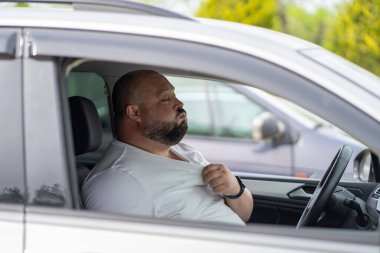 Yorgun adam, arabanın içinde sıcak havada kan basıncını hissediyor. Aşırı ısınma nedeniyle trafik sıkışıklığında araba süren aşırı kilolu erkek, pencereler açık serinlemeye çalışırken durur.