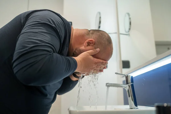 stock image Overweight balded middle aged man washing face in bathroom at home under sink splashing water, side view. Daily routine, hygienic procedure, morning treatment and cleanliness of body concept.