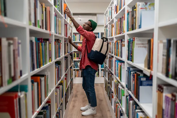 大学図書館で本を選ぶアフリカ系アメリカ人のオタク学生 大学を勉強バックパック付きの賢明な男ヒップスター 新しい職業の提供 学習コンセプト — ストック写真