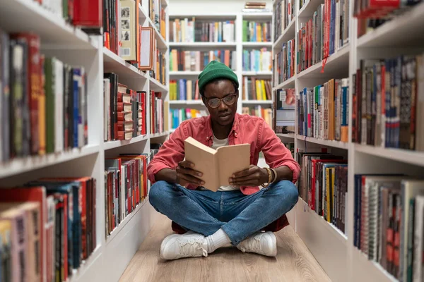 学生アフリカ系アメリカ人の男性は 本棚の間の床に座って大学図書館で研究教科書を読んでいる 公共の場所で勉強する気の利いた男ヒップスター — ストック写真