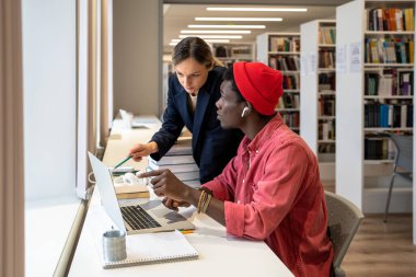 Concentrated diverse couple of students working on laptop together on study project in university library. Serious college teacher pointing out on computer screen mistake in solution to student guy clipart