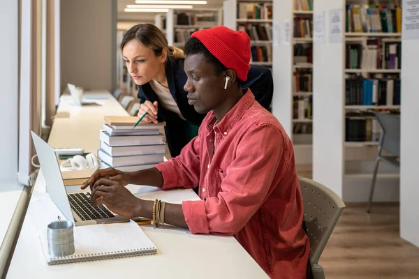 興味のある集中アフリカ系アメリカ人の学生の男性は 深刻な大学の家庭教師の女性の近くのノートパソコンを入力する画面を鉛筆を保持 試験準備プロジェクトに取り組む多様なカップルに焦点を当て — ストック写真