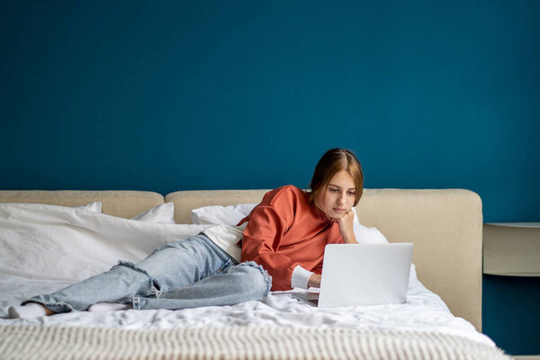 Сосредоточенная девочка-подросток, работающая над учебным проектом на ноутбуке, лежит дома и смотрит на экран. Внимательный серьезный кавказский подросток печатает текст. Обучение, изучение, поиск информации на компьютере.