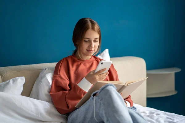 10代の少女が自宅のベッドに座っているソーシャルメディアで共有するためにスマートフォンでページを撮影本を読んでいます 白人のティーンエイジャーの本ワームオタク文学の種類 レジャー 携帯電話で写真を撮る — ストック写真