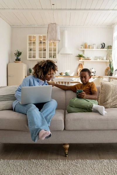 African american mom working on laptop and looking at smartphone in sons hands sitting on couch at home. Teen boy browsing in smartphone, watching videos. Working busy mom, child gadget addiction.