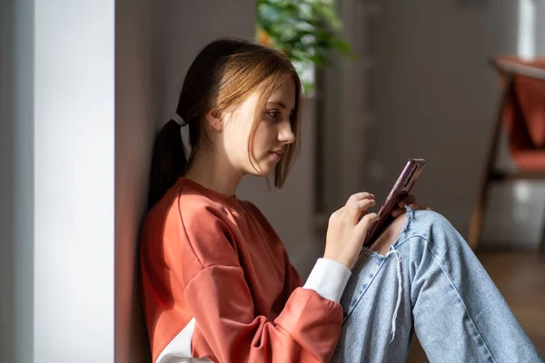 严肃的少女坐在家里的地板上 边看边用智能手机在社交媒体上浏览 青少年与朋友聊天 看屏幕 在网上用手机上网 — 图库照片