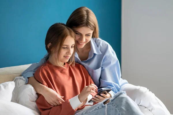 积极的妈妈女儿一起坐在床上看智能手机里有趣的视频 家庭妇女少女浏览手机搜索信息 数码娱乐 — 图库照片