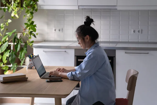焦点を当てたアジアの女性のフリーランスは キッチンの自宅にラップトップ付きのテーブルにリモートで座って働いています 自給自足の若い韓国の女性のコピーライターは雑誌または自身の本のための記事をタイプしている — ストック写真