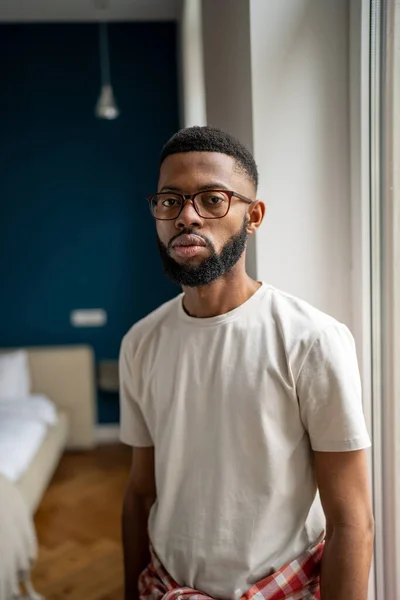 Serious calm arfrican american bearded guy in eyeglasses standing near window at home. Isolated thoughtful young black man freelancer wearing simple white t-shirt looking at camera in living room.