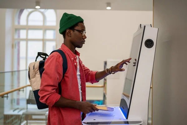学生アフリカ人の男性は セルフサービスの電子端末を使用して 営業担当者なしで店で商品を支払う タッチスクリーン自動販売機を使用して大学で屋内に立つ自給自足の黒人男性 — ストック写真
