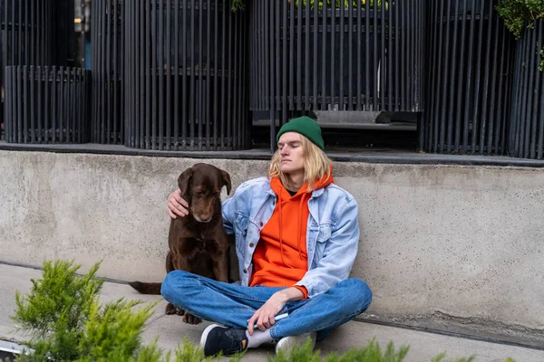 Upset depressed millennial guy sitting on sidewalk with dog, having housing problems. Lonely young man on city street thinking about life problems. Homelessness and mental health