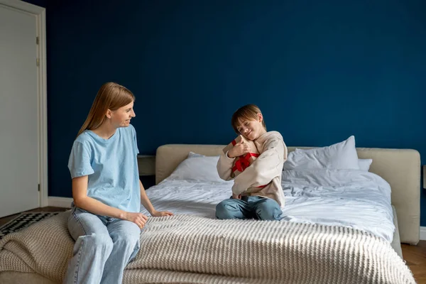 stock image Grateful little son hug wrapped gift box sits on bed. Caring loving mother greeting child with birthday. Happy family celebrating holiday together at home. Kid boy smiling and excited holding present 