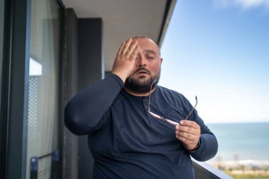 Suffering overweight man feeling headache rubbing eyes stands on balcony on hot weather in summer. Sweating tired guy suffer pressure problems living in tropical climate high humidity. Health problems clipart