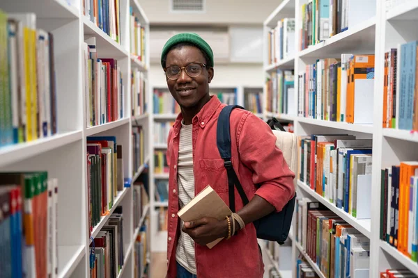 微笑着站在书架之间的非洲裔美国学生在大学图书馆里拿着书 校园生活 自学书籍学习学习研究理念 — 图库照片