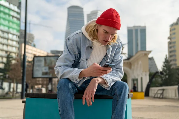 Worried guy hipster looking in phone sitting on bench in city with modern cityscape on background. Serious puzzled man has bad news browsing internet, surfing online, reading message on smartphone.
