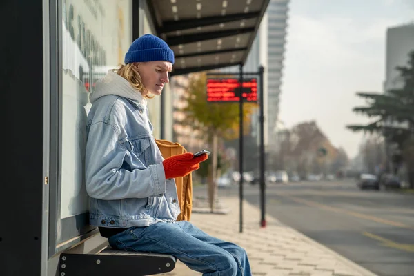 白种人千禧年学生清晨坐在巴士站的长椅上 手持智能手机 等待交通 在线查看校车时刻表 城市公共交通基础设施 — 图库照片