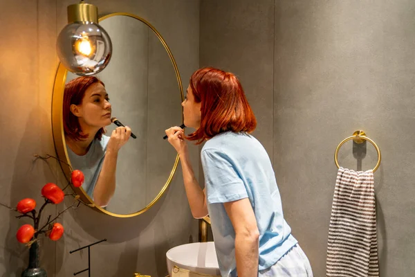 ブラシで顔に毎日メイクを施した鏡を見ている赤毛の女の子 若い女性が欠陥をカバーするためにコンパクトな粉を使用してバスルームに立っている 社会の美しさの基準に適合 — ストック写真