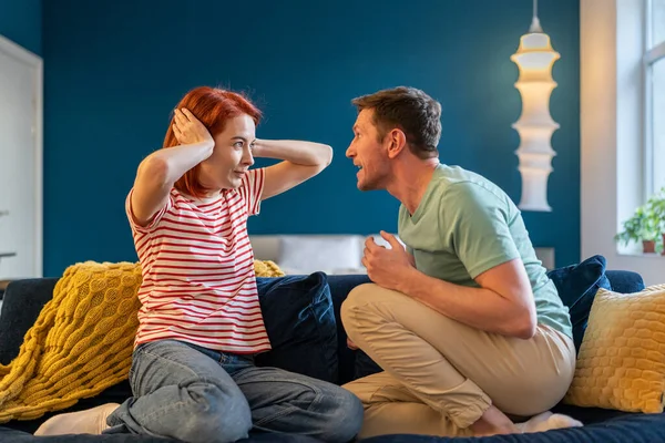 Angry annoyed man husband screaming at wife covering ears with hands, Caucasian couple sitting together on sofa and arguing fighting at home, having misunderstandings in relationship. Emotional abuse