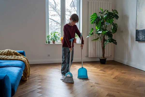 子供の家事 子供が床を掃除してる 家で彼の部屋を掃除しながら掃除する方法を学ぶ 家をきれいにするのを助けるほうきとシャベルを持っている子供の男の子 — ストック写真